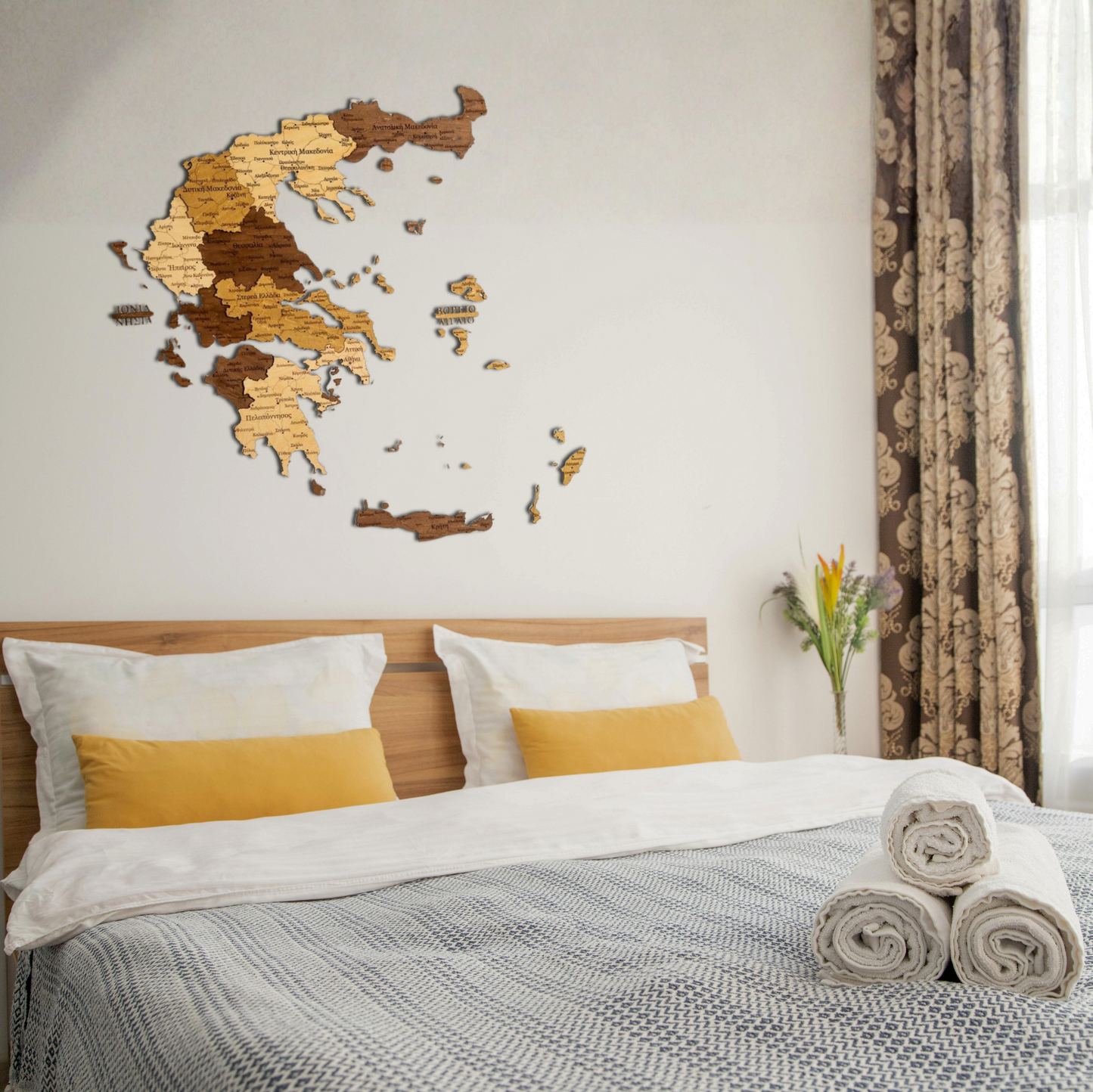 Bedroom with wooden wall art of a Greece map in Greek language, bed with white and yellow bedding, and side table with flowers.