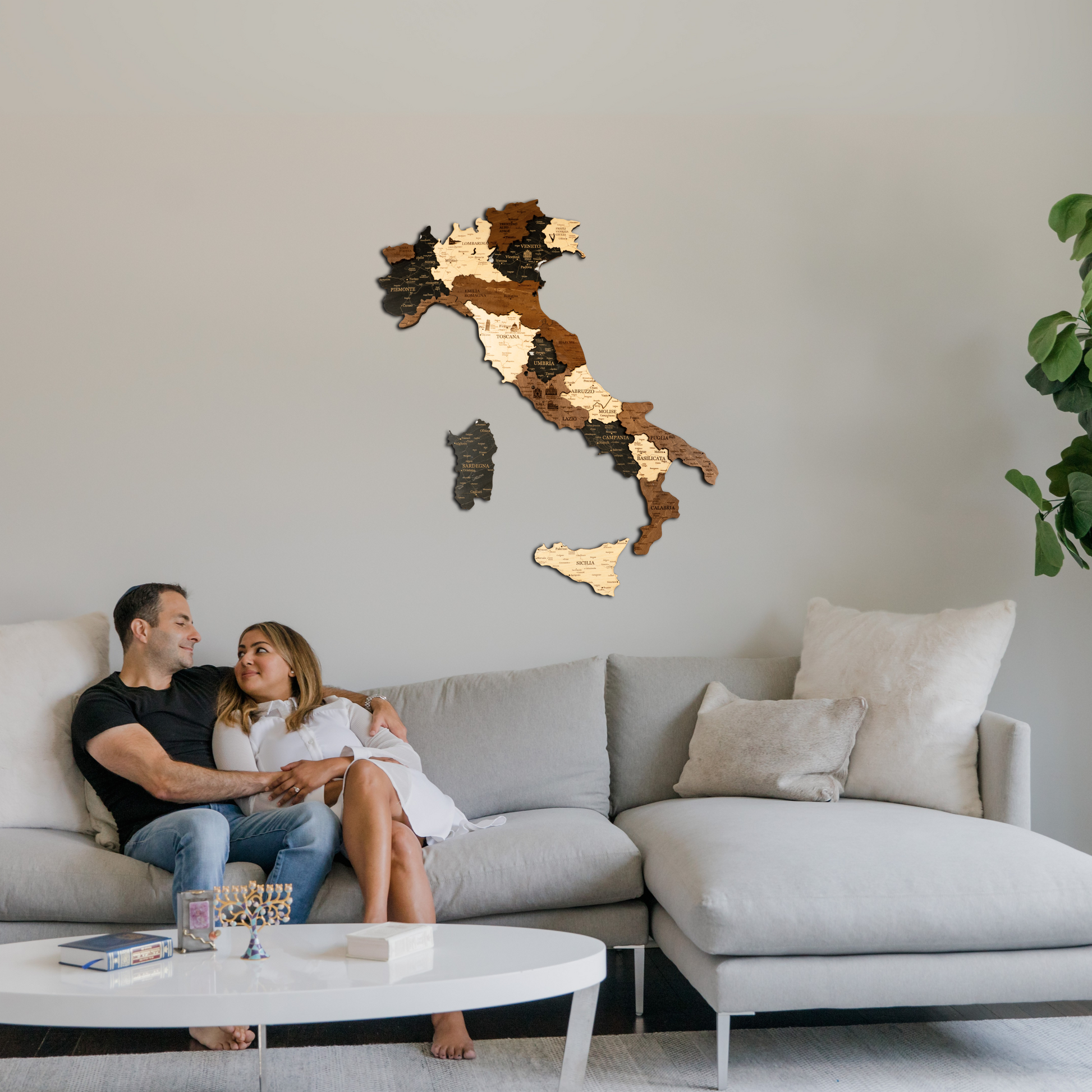 Couple sitting on a couch in a living room with a wooden map of Italy on the wall.