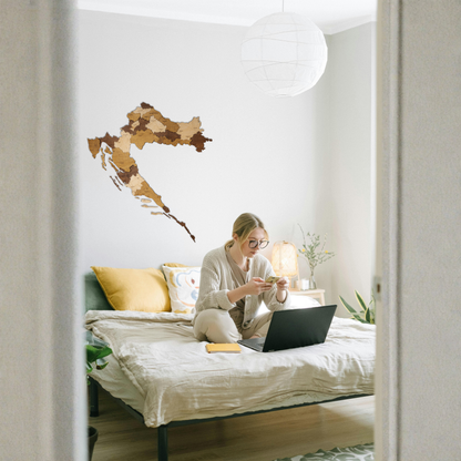 Woman sitting on a bed using a laptop with a decorative wall map of Croatia in the background