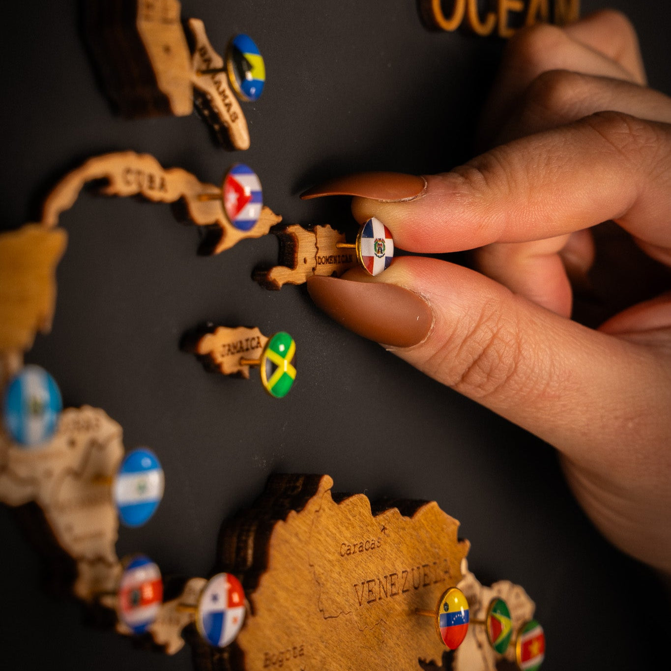 A person's hand placing a push pin with a national flag on a wooden map of South America.